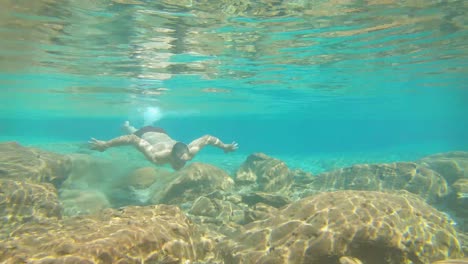 Joven-Nadando-En-Azul-Claro-Bajo-El-Agua-De-La-Cascada-Con-Muchas-Rocas-Desde-Un-ángulo-Plano