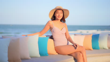 An-attractive-woman,-in-her-swimming-suit-and-a-sun-hat,-sitting-on-an-outside-bench-with-the-ocean-in-the-background