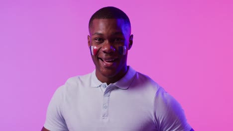 Portrait-of-happy-african-american-male-football-supporter-with-flag-of-france-over-pink-lighting