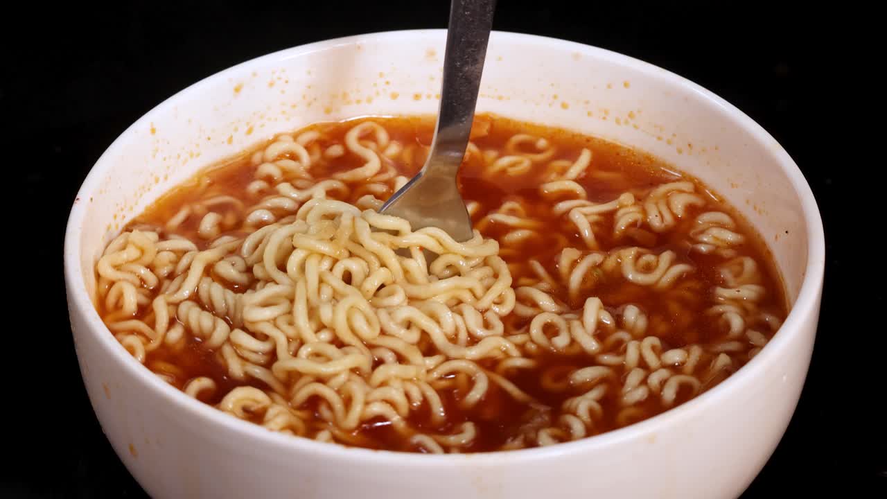 A metal fork twirls and lifts cooked instant noodles from a white bowl of red broth, under bright studio lighting with a black background