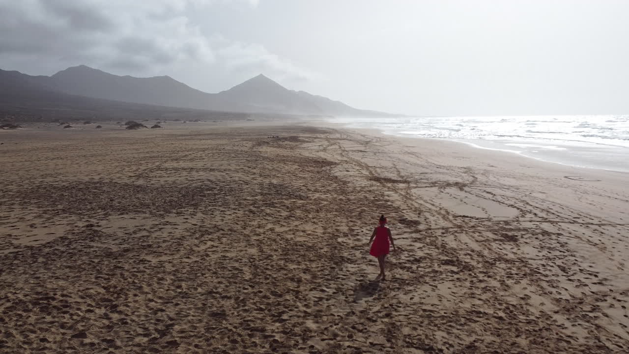 A woman in red dress walking alone on a sunny beach in Fuerteventura