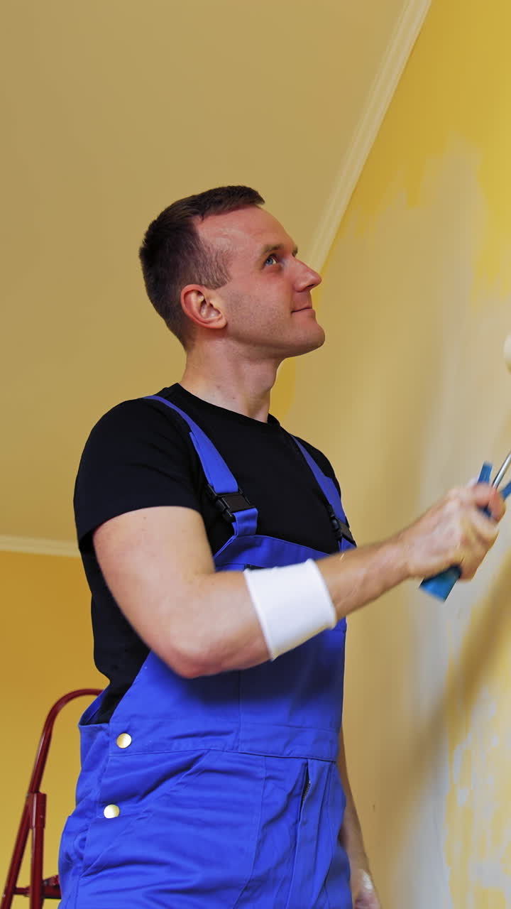 Funny craftsman painting walls. Cheerful young male worker imitating singing on a paint roller during his work on room renovation. Vertical video
