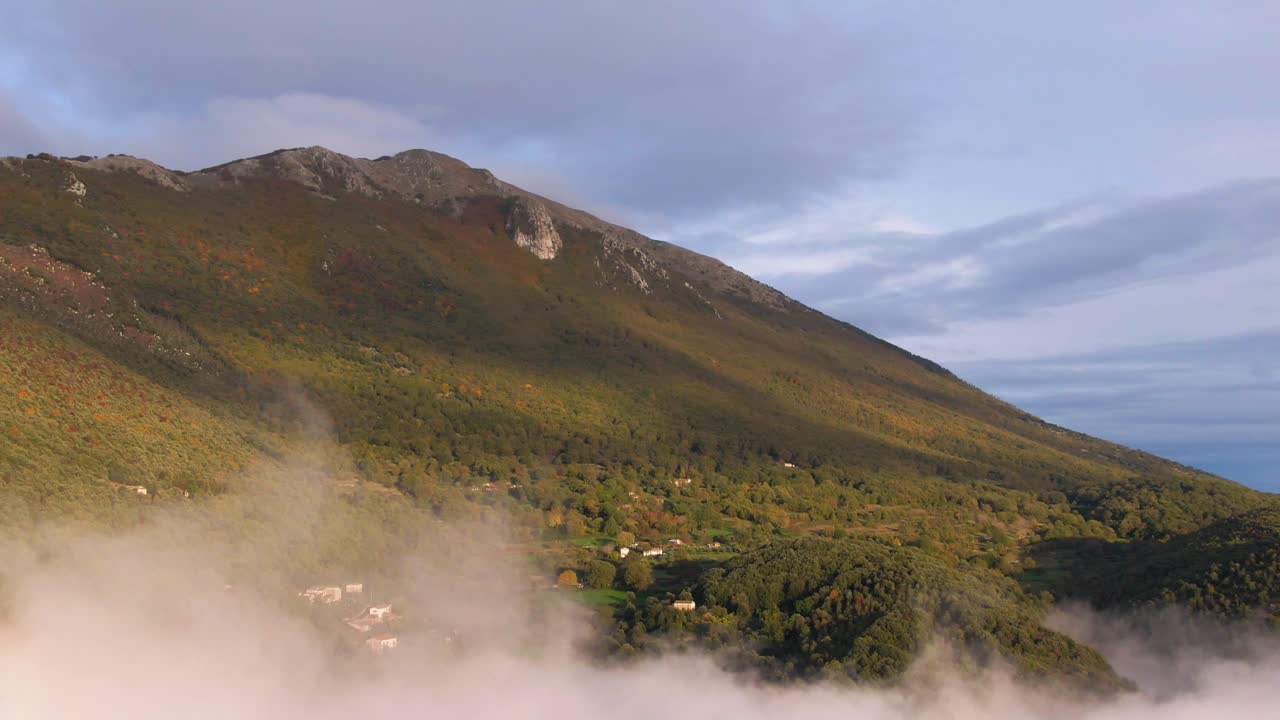 Flight through opaque clouds to reveal green mountain and buildings nestled beneath, aerial approach