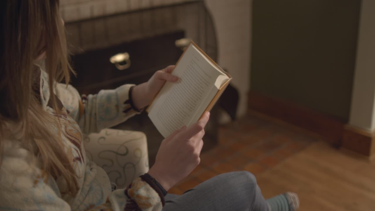 A close up over the shoulder shot of a woman in a sweater reading a book, closing it, and putting it down. In a vintage cozy living room. 60 frames for slow motion