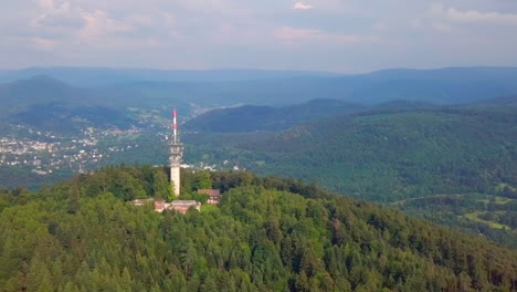 approaching aerial view of the 'fremersberg' transmission tower on a green forested mountain, revealing the spa town of baden-baden in the background on a sunny summer day.