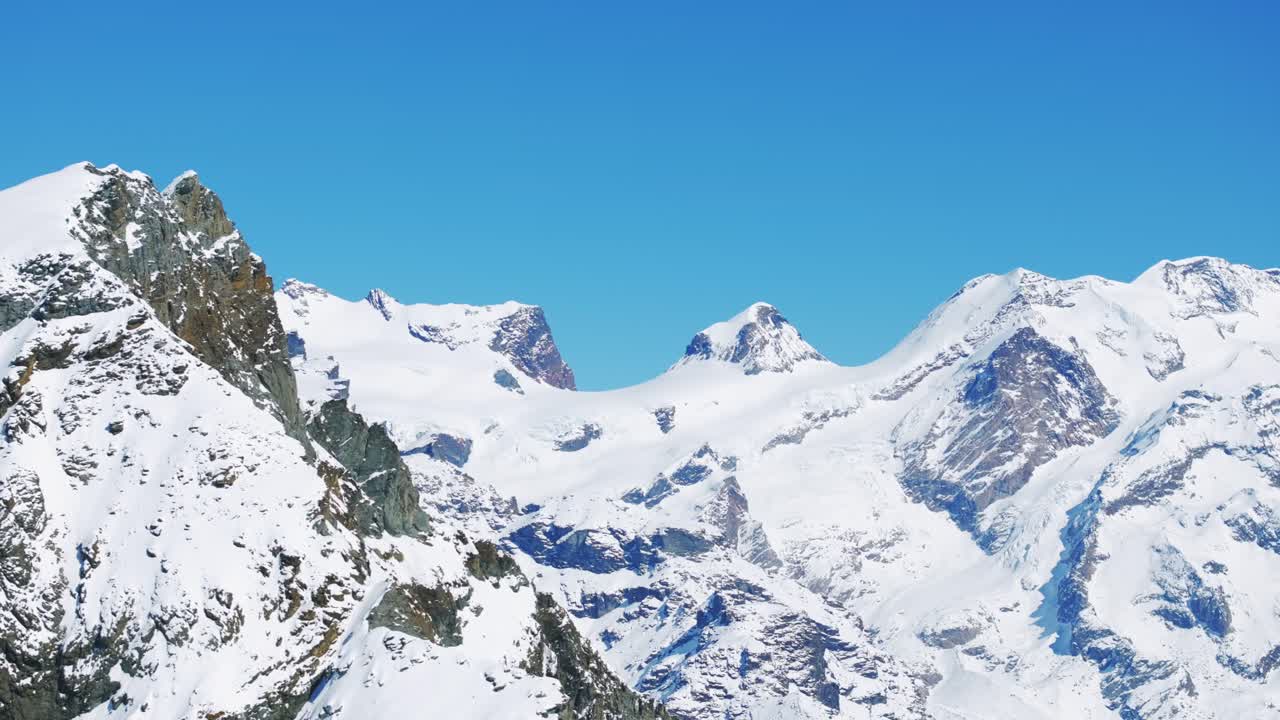 Sweeping panoramic view of vast snow-covered Alpine mountain range under bright blue sky, ideal for travel, nature documentaries, winter scenes. Aerial lateral view, copy space