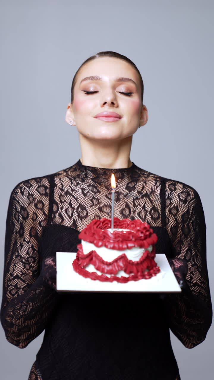 Beautiful Caucasian model wearing long-sleeve black lace suit holding a cake. Woman makes a wish and blows the candle. Vertical video.