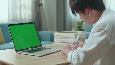 Close Up Of Asian Man Student Look At Green Screen Computer And Write In The Notebook While Studying Online At Home
