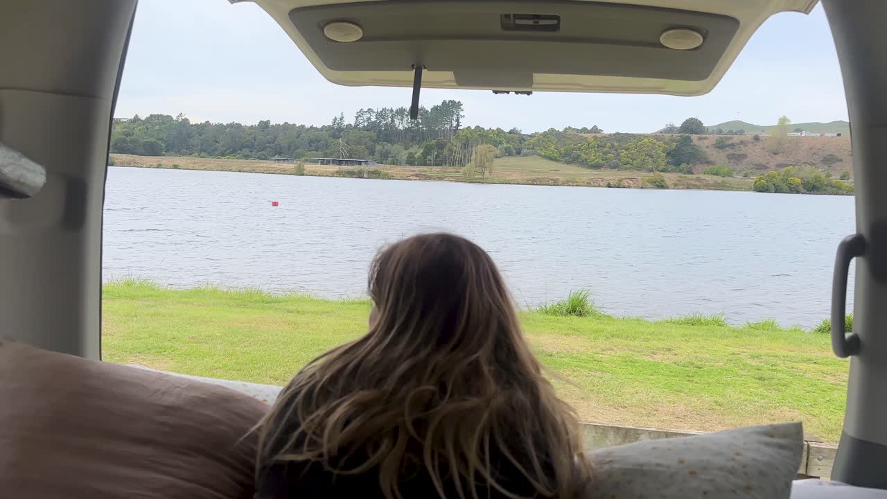 A girl looks out from the back of her camper van towards a serene lake at Horahora Domain Camp, New Zealand. Calm waters, lush greenery, and peaceful surroundings create a tranquil scene
