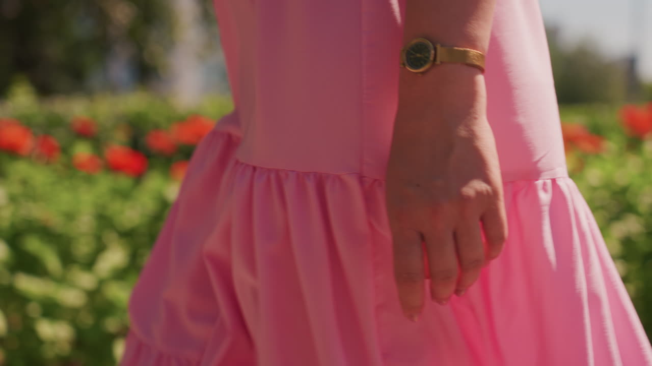 primo piano di una mano con un abito rosa che passeggia in un giardino soleggiato con fiori rossi, orologio che luccica al polso, dita che sfiorano le increspature, delicato tepore estivo, elegante slow motion, musa fashion blogger