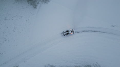 Snow-plowing-tractor-with-glowing-lights-in-winter-landscape,-aerial-top-down-view