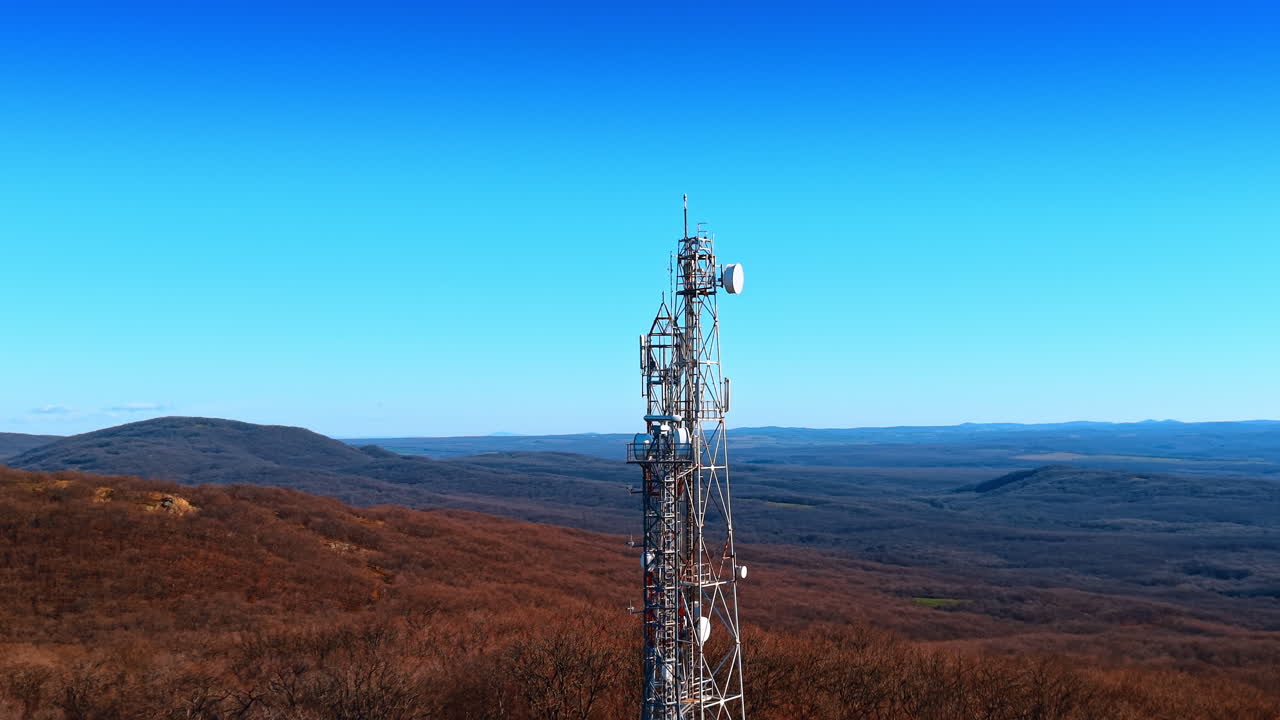 Torre per telecomunicazioni su una collina boscosa sotto un cielo azzurro e sereno. Traliccio metallico con antenne e parabole satellitari su una dorsale boscosa con colline ondulate.