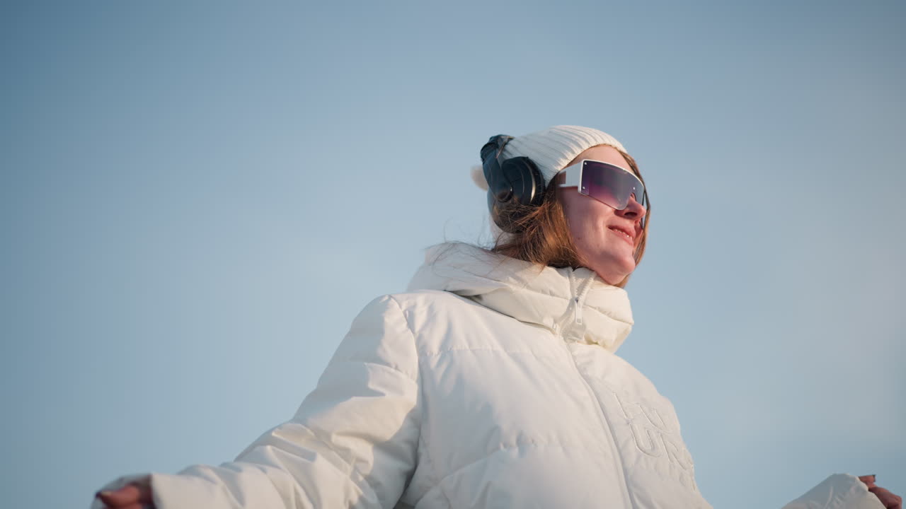 Young artist smiling joyfully under sunlight while grooving in large headphones and sunglasses, dressed in white winter jacket, enjoying warmth of winter day with blurred cityscape