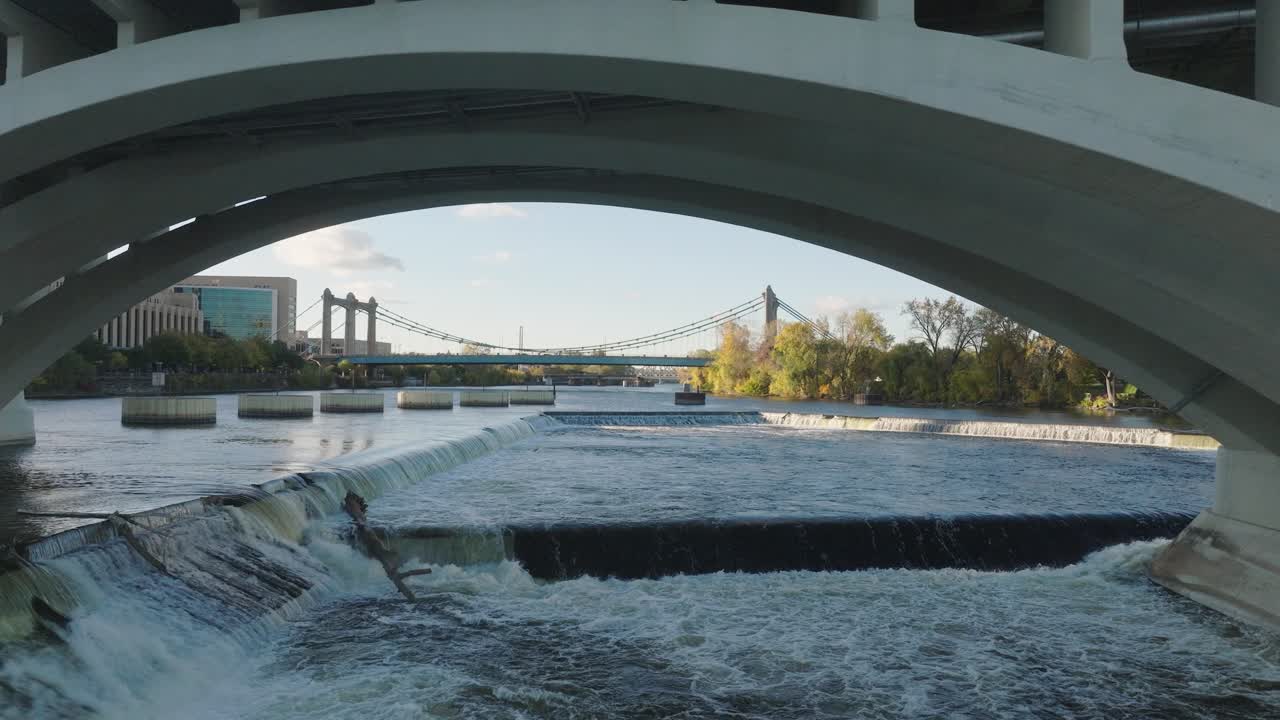 Low aerial dolly shot under the Third Avenue Bridge arch, revealing the St. Anthony Falls Dam spillway and the blue Hennepin Avenue Bridge at golden hour