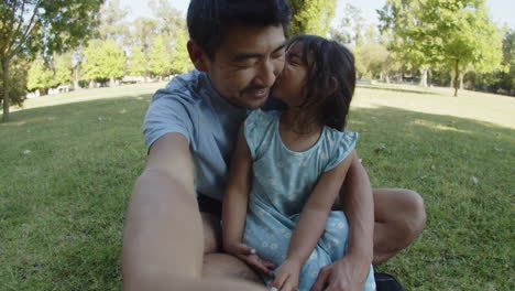 Glücklicher-Vater,-Der-Mit-Tochter-Auf-Gras-Sitzt-Und-Ein-Video-Macht,-Während-Das-Mädchen-Ihn-Küsst