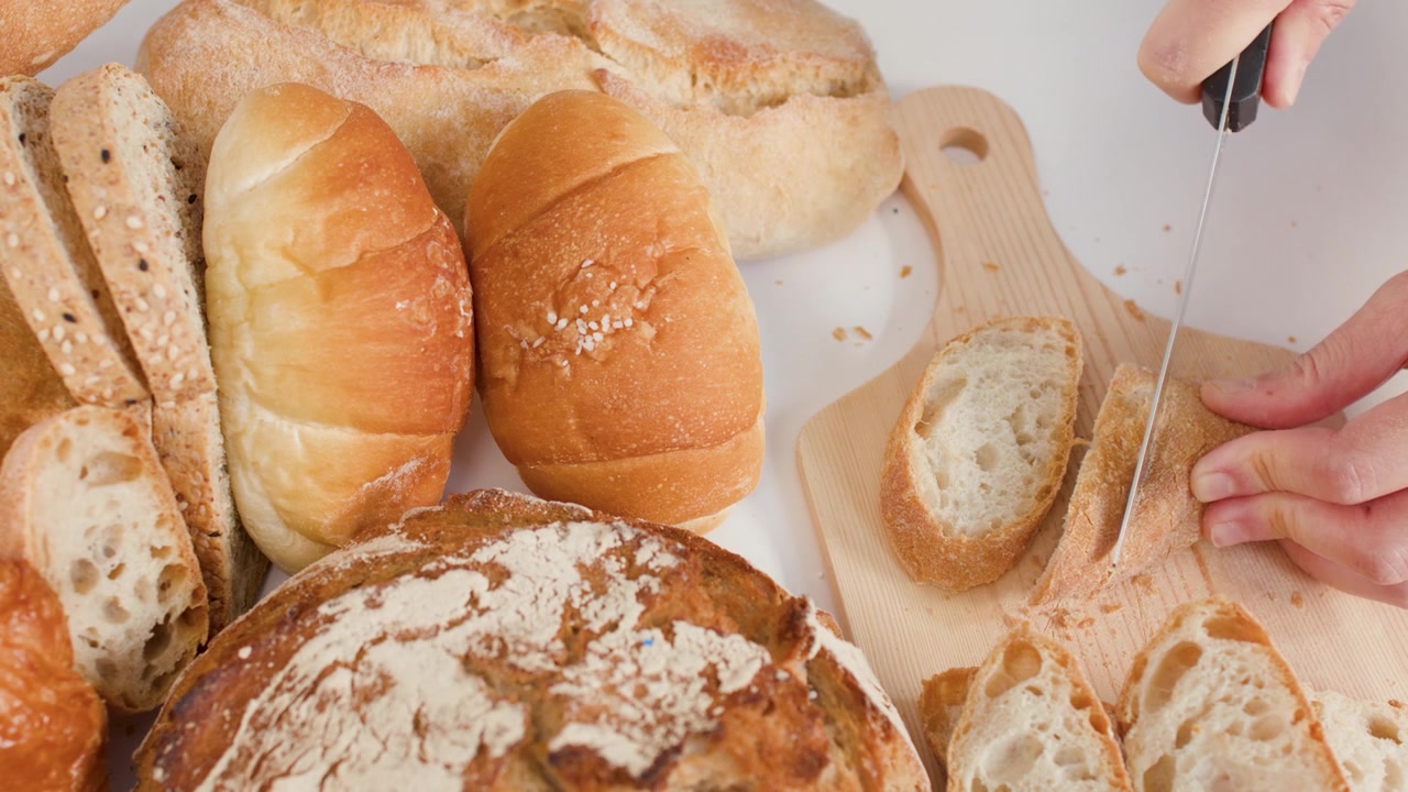 Close-up of hands slicing crusty bread on a wooden board amidst various artisan loaves.