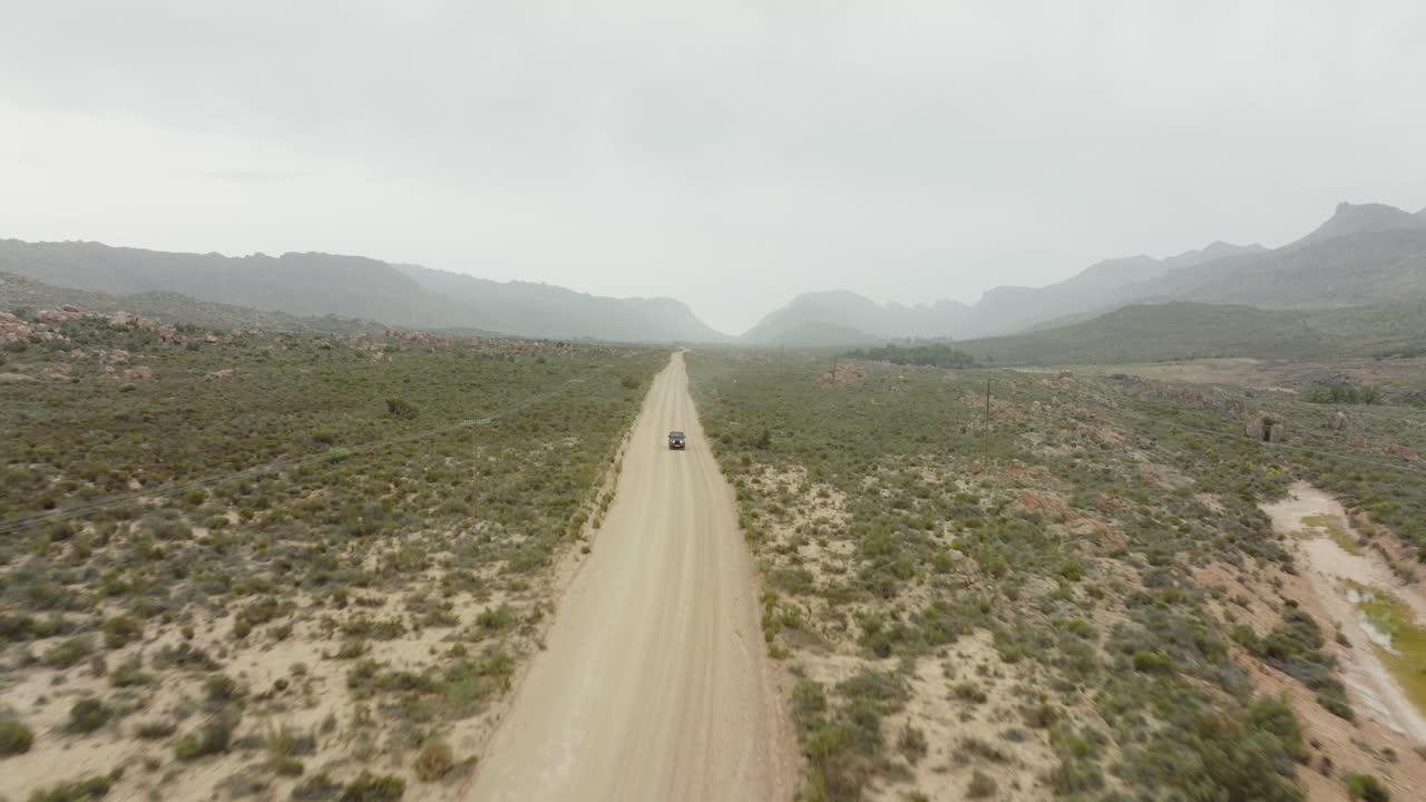 el avión no tripulado vuela hacia atrás sobre una carretera y sigue un vehículo fuera de carretera negro a vista de pájaro - paisaje desértico en el área silvestre de cederberg en sudáfrica - se pueden ver montañas en el fondo