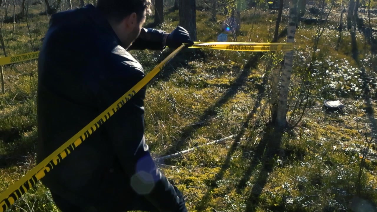Vista posteriore di un agente di polizia con giacca di pelle e guanti che si china sotto un nastro giallo di delimitazione "Scena del crimine non oltrepassare" per entrare in un'area di indagine riservata in una foresta soleggiata