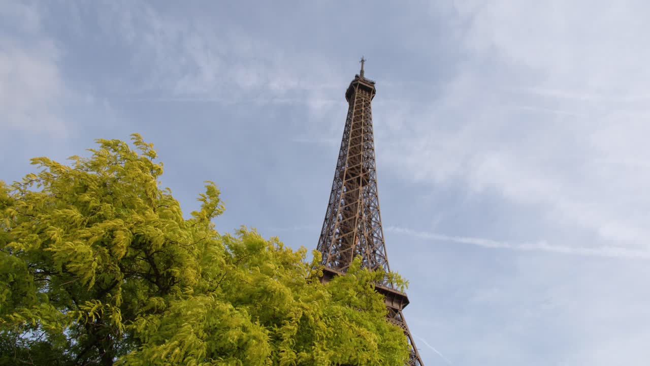 Inquadratura statica dal basso della cima della Torre Eiffel incorniciata da alberi estivi di un verde vibrante