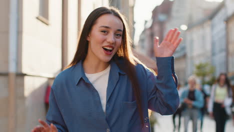 mujer joven sonriendo amigablemente a la cámara, agitando las manos hola, hola, saludo o adiós en la calle de la ciudad