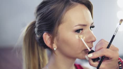 primer plano de un artista de maquillaje profesional aplicando corrector a la piel de una mujer joven con pincel para cubrir las ojeras y maquillar la cara