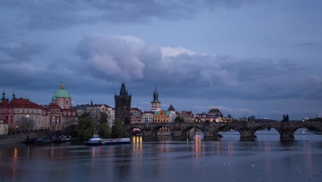 Day-to-night-sunset-timelapse-along-the-Charles-Bridge-on-the-Vltava-river-with-boats-on-a-cloudy-evening-in-Prague,-Czech-Republic-as-the-bridge-and-towers-of-the-city-light-up