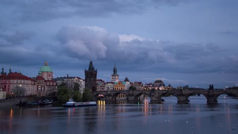 day to night sunset timelapse along the charles bridge on the vltava river with boats on a cloudy evening in prague, czech republic as the bridge and towers of the city light up