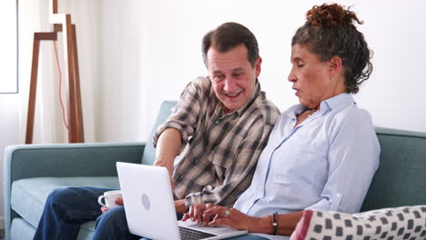 Senior-Couple-Sitting-On-Sofa-At-Home-Using-Laptop-Computer-To-Shop-Online