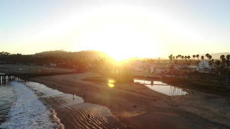 Aerial-drone-shot-at-sunset-rising-up-over-the-pacific-ocean-beaches-while-looking-over-the-palm-trees-at-the-beautiful-city-of-Santa-Barbara,-California