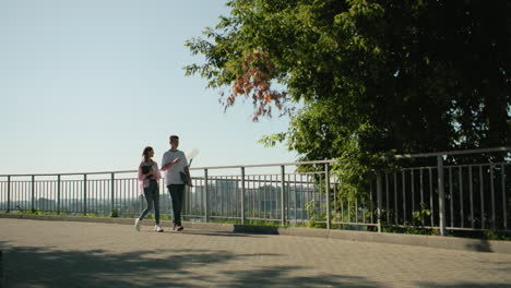 students walking on paved path near iron railing holding books, engaging in cheerful conversation surrounded by greenery, cityscape, and urban architecture under bright sunlight