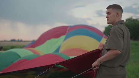 man holding colorful balloon envelope sections by braided cables during preflight preparation in open green field at dawn under soft pastel sky with sunrise glow reflecting on fabric folds