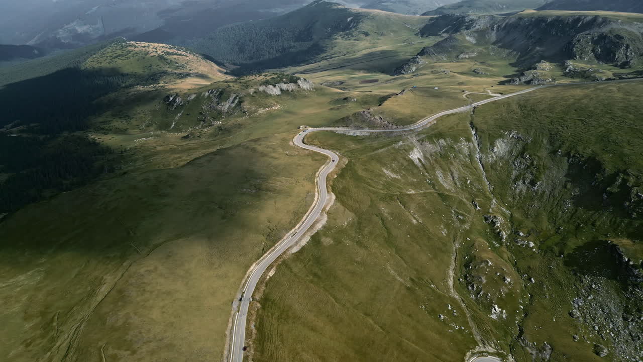 perspectiva aérea de la carretera transalpina en rumania, revelando su camino serpenteante a través de terrenos verdes exuberantes, bosques sombreados y bordes montañosos escarpados