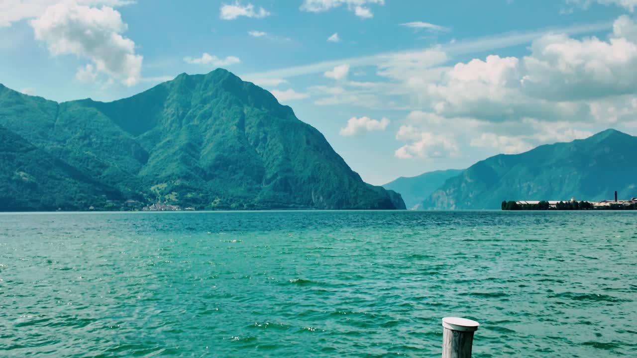Vista dalla ringhiera di un pontile metallico che si affaccia su acque blu profonde verso le ripide montagne del Lago d'Iseo. Giornata di sole in una località alternativa al Lago di Como che mostra un orizzonte limpido e le Prealpi italiane.