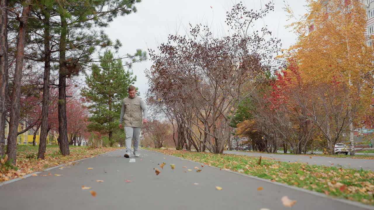 Autumn foliage flows with wind as boy walks on tarred path in urban park surrounded by colorful trees and fallen leaves, with buildings and cars in background on cool overcast day with breeze