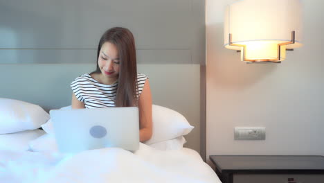 a young woman working on her laptop in the comfort of a hotel suite bed reacts to what she sees on the screen with a nonverbal sign of triumph