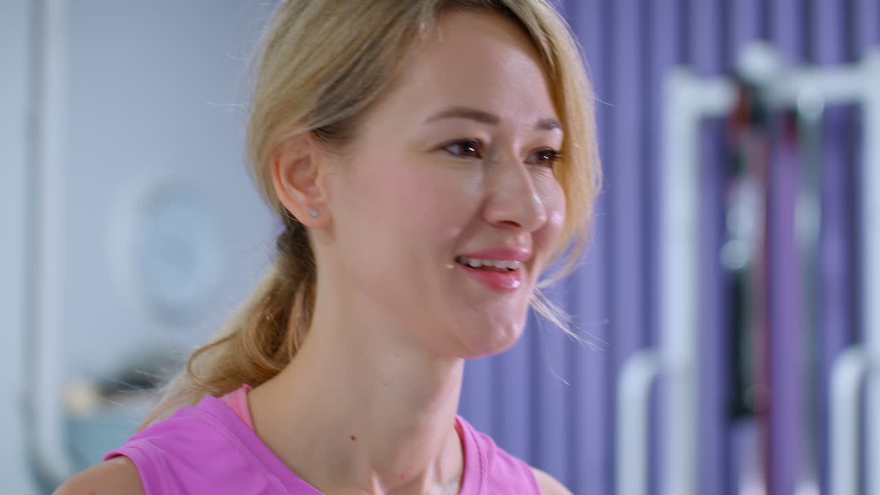 핑크색 옷을 입고 머리카락을 뒤로 은 미소 짓는 여자의 클로즈업, 운동 운동을 하는 동안 대화에 참여, 그녀는 몸을 구부리고 체육관 환경에서 일어납니다.