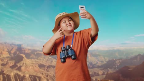 asian boy with a hat and binoculars taking a selfie on smartphone while traveling at the top of mountain. boy researcher examines something, travel tourism adventure concept