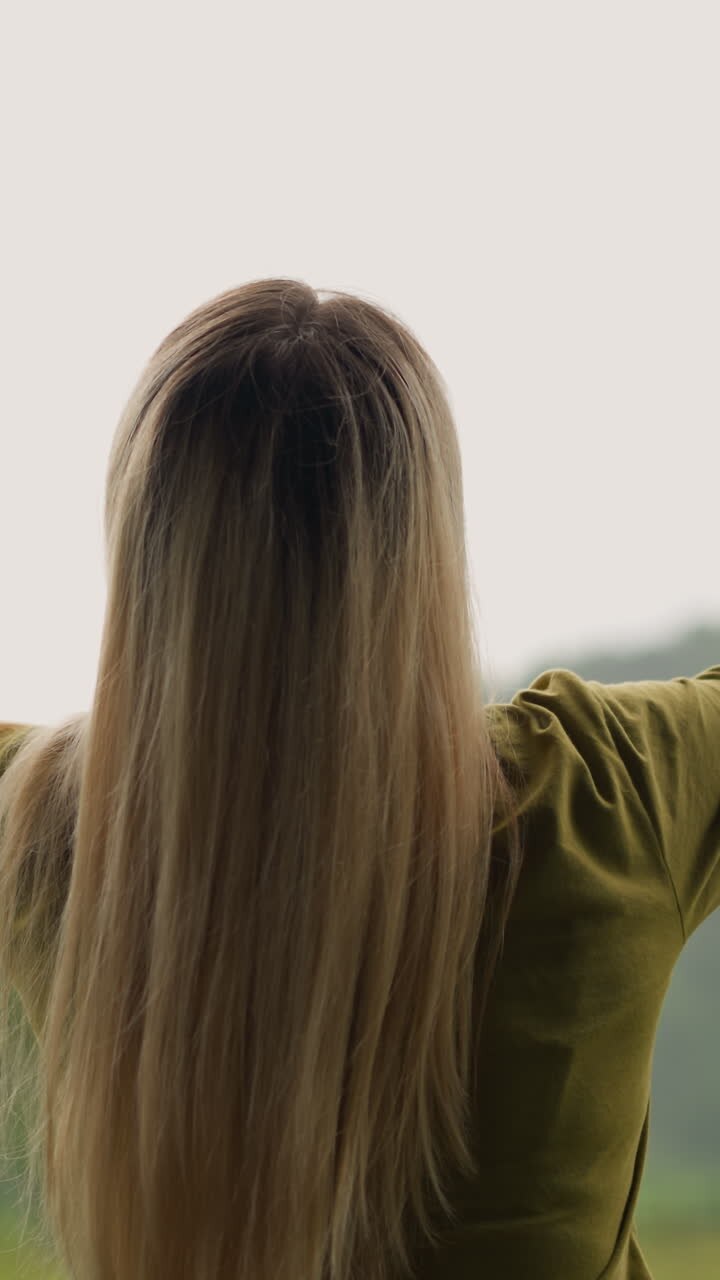 Cheerful young woman raise hands up happy of warm rain against green sloppy hill at eco resort on nasty day backside view slow motion