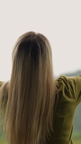 cheerful young woman raise hands up happy of warm rain against green sloppy hill at eco resort on nasty day backside view slow motion