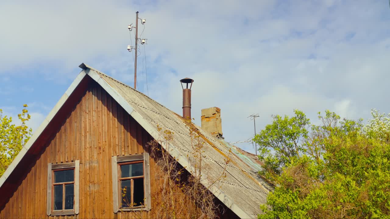 Rustic wooden house with chimney and utility pole visible above a weathered roof, surrounded by spring trees. Daugavpils, Latvia (Latgale)