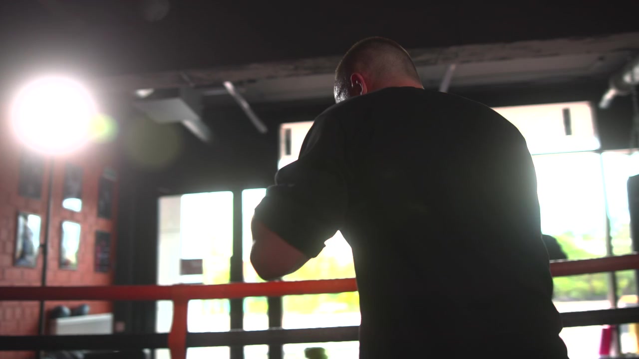 Un pugile adulto si trova in una palestra di allenamento poco illuminata accanto alle corde rosse del ring, con i pugni alzati vicino al viso, le spalle tese, il sudore visibile, con un forte controluce, ombre intense e un'atmosfera di preparazione concentrata.