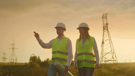 An-electrician-male-and-female-in-the-fields-near-the-power-transmission-line.-He-is-an-electrician-who-manages-the-process-of-erecting-power-lines.-The-mechanic-in-a-helmet-and-Manager-with-tablet