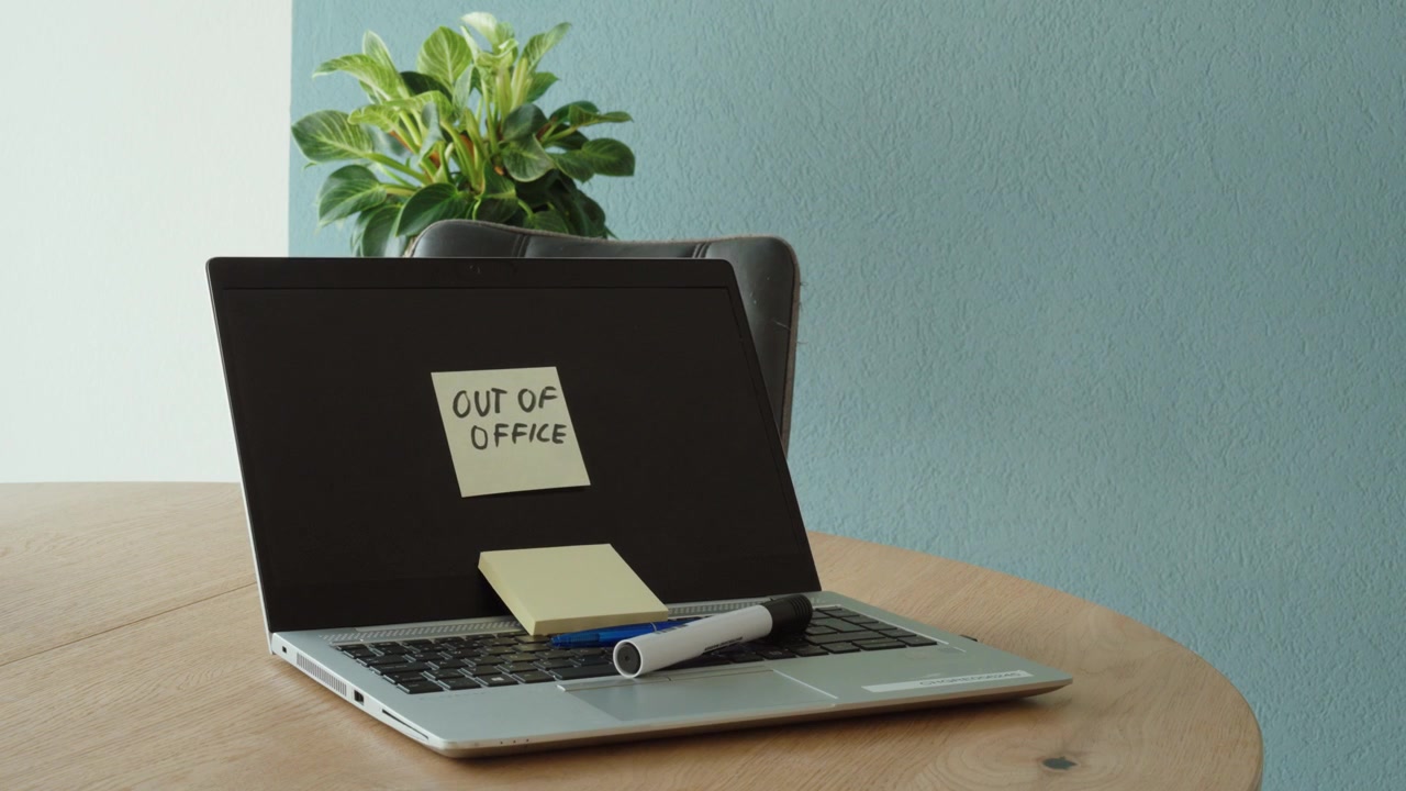 A person places an "Out of Office" sticky note on a laptop before leaving the workspace. Concept of vacation, time off, workplace communication, and temporary absence.