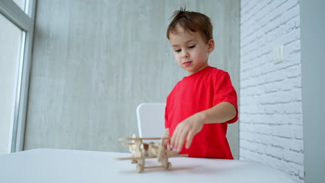 beautiful little kid in red t-shirt stands at the desk. lovely baby touches a wooden toy airplane. low angle view.