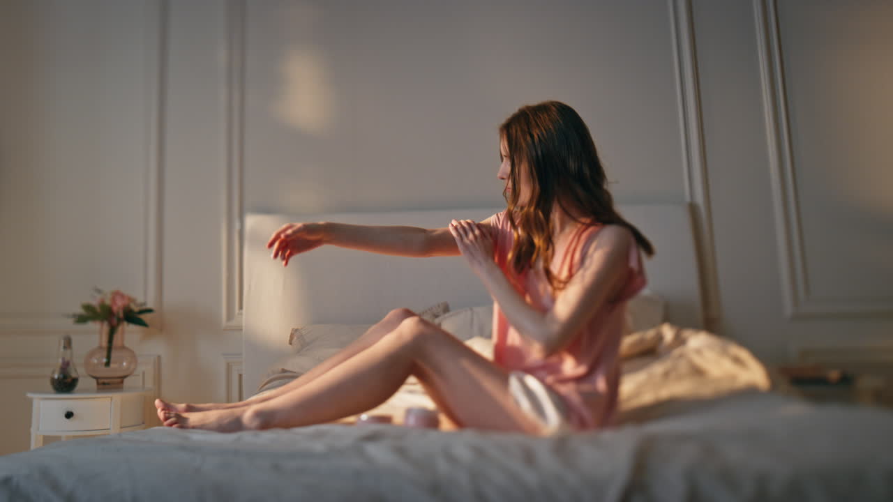 매력적인 여성이 쾌적한 빛 침실에서 로션을 적용합니다. 편안한 소녀 마사지