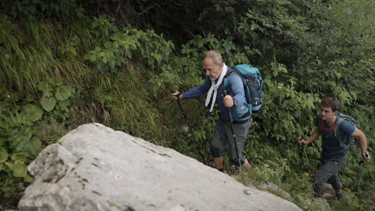seguimiento de cámara en frente, dos excursionistas caminando por una colina en un camino estrecho cerca de un bosque verde, camino lleno de vegetación, hojas y otras plantas