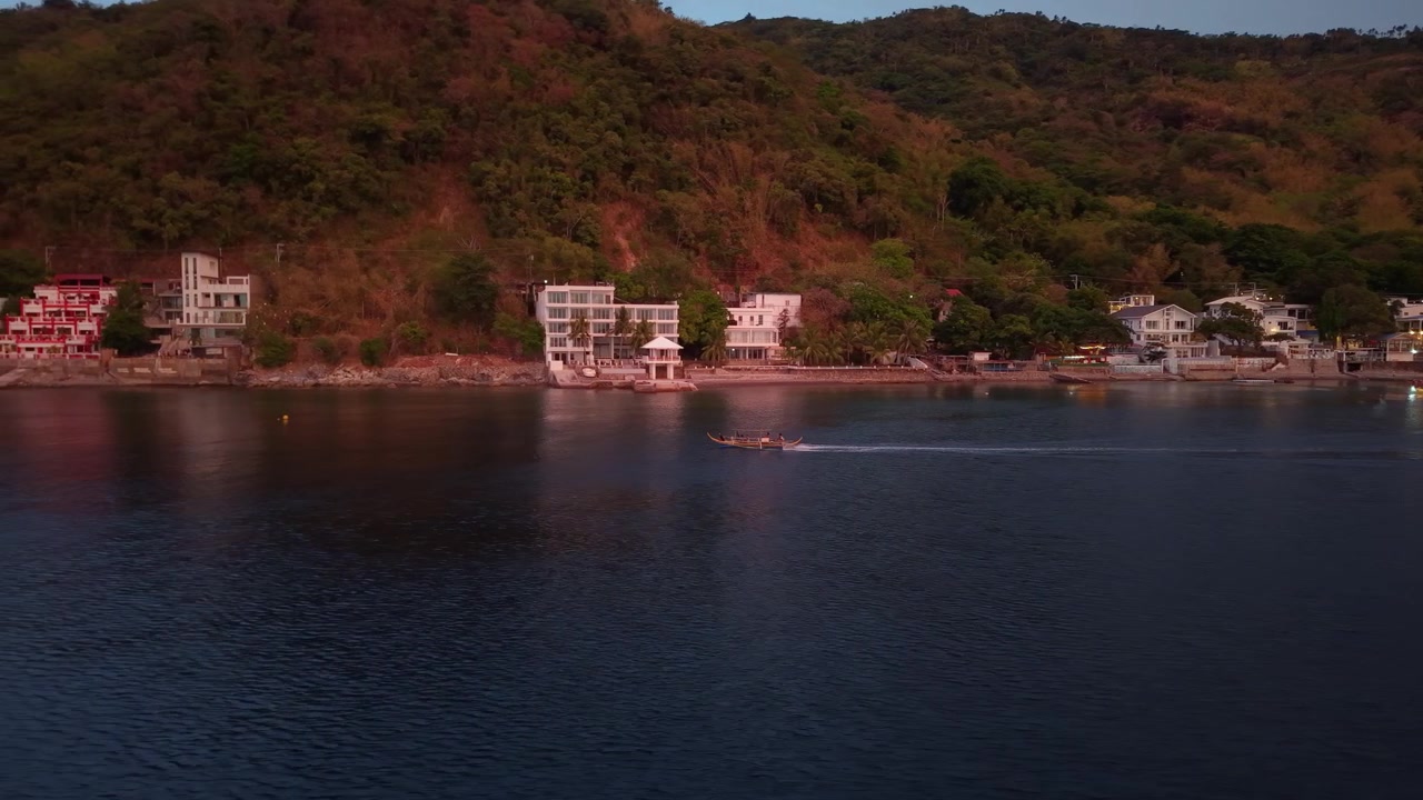 Riprese aeree con drone di una barca da pesca solitaria che naviga su acque tropicali calme al tramonto di fronte alla costa di Anilao, creando un paesaggio marino pacifico e suggestivo.