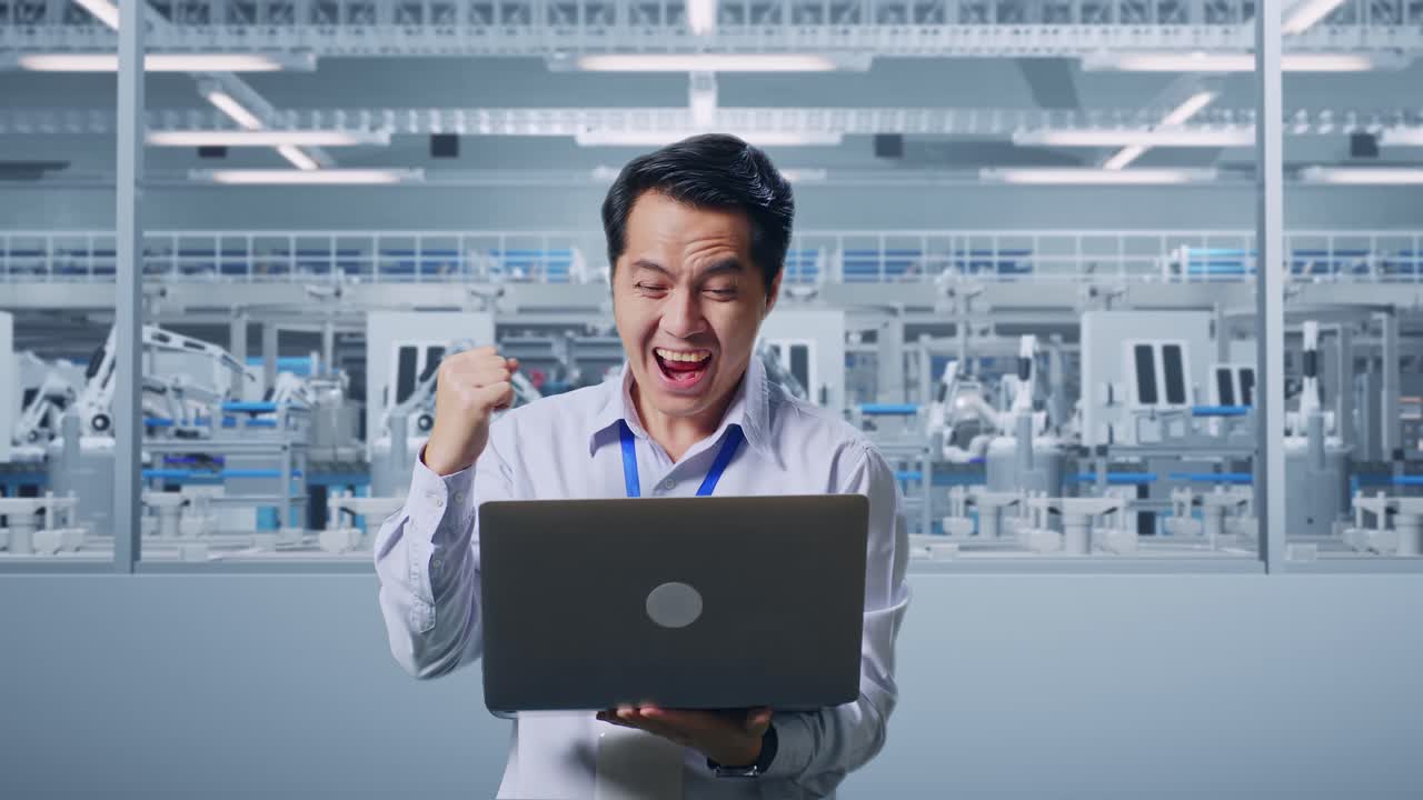Asian Male Professional Worker Standing With His Laptop At Factory Digitalization, He Raises His Fist Up With Screaming Goal