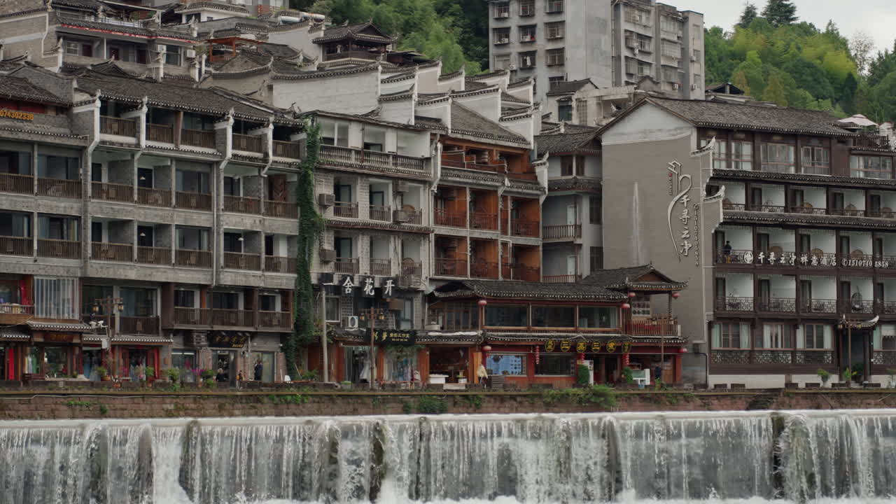Edifici storici sul lungofiume accanto a una cascata, facciate in pietra invecchiata e salici, acqua che scende a cascata su una piccola diga, scalinate lungo il fiume e vicoli stretti, piccoli negozi e terrazze, superficie riflettente.
