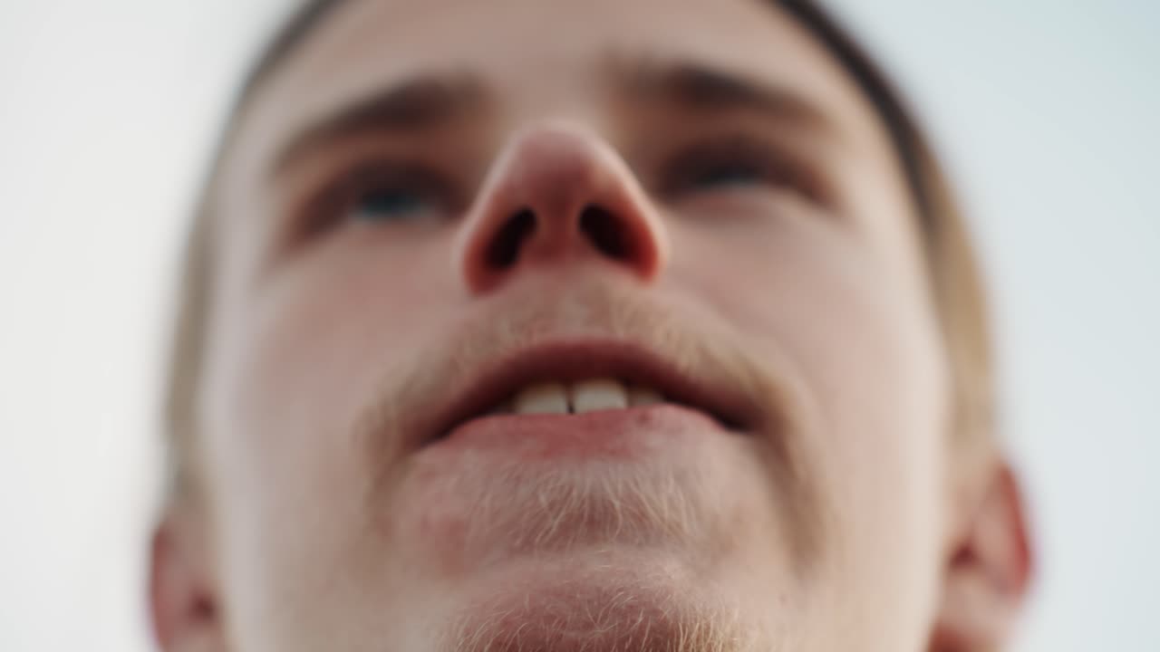 Primo piano dal basso di un uomo bianco con la mascella serrata, labbra contratte, sguardo verso l'alto nel cielo pallido, leggera barba incolta, tensione che si trasforma in un leggero sorriso, scatto eroico all'aperto, atmosfera urbana, pronto all'azione.